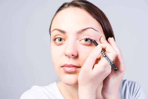 Close up Smiling Young Woman Applying Eyeliner Makeup to Left Eye While Looking at the Camera on a Gray Background.